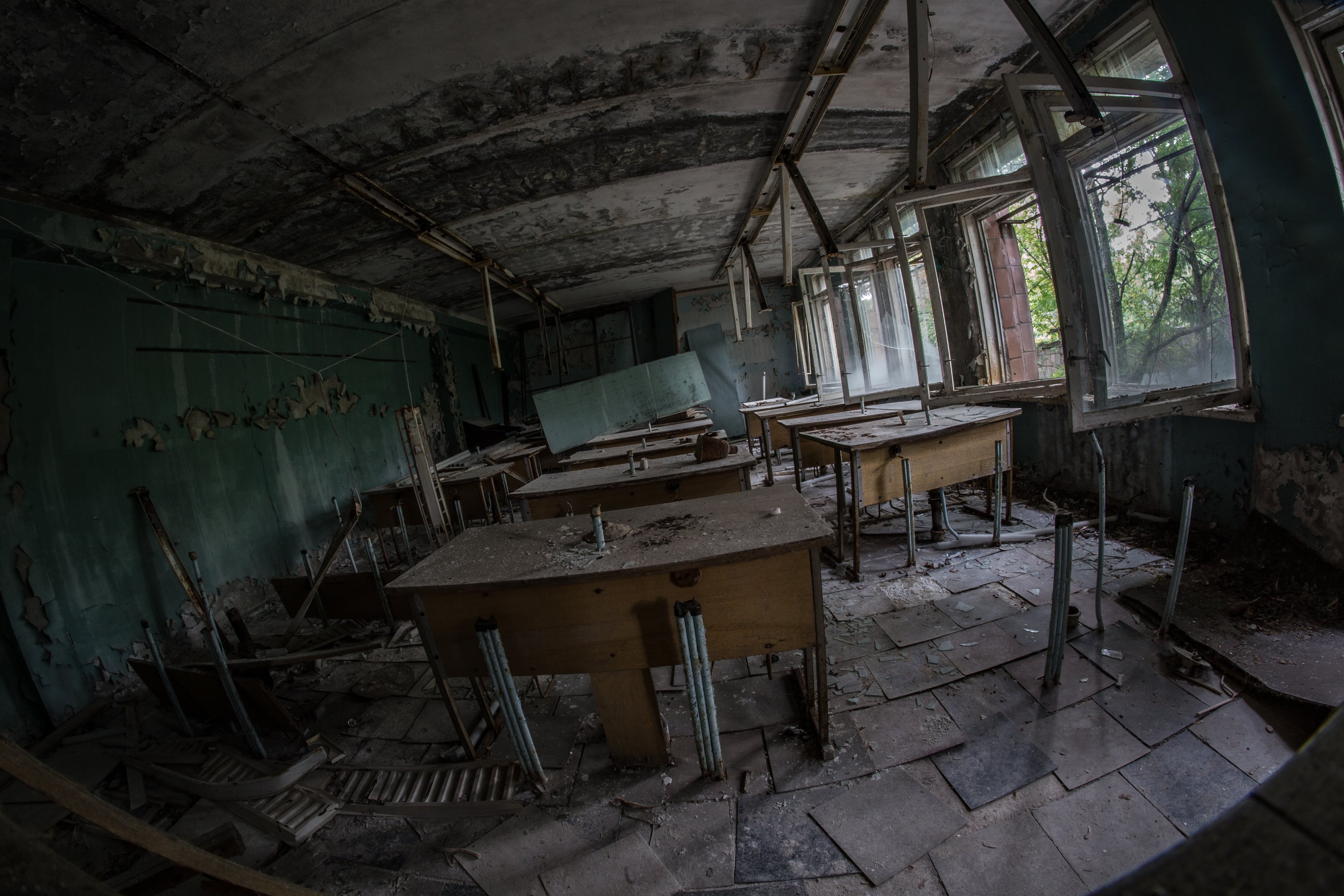 Verlassener Klassenraum in Pripjat, Tschernobyl, Ukraine, mit Tischen, Stühlen, abblätternder Wandfarbe, kaputten Fenstern und sichtbarer Decke, Bäume durch Fenster zu sehen.