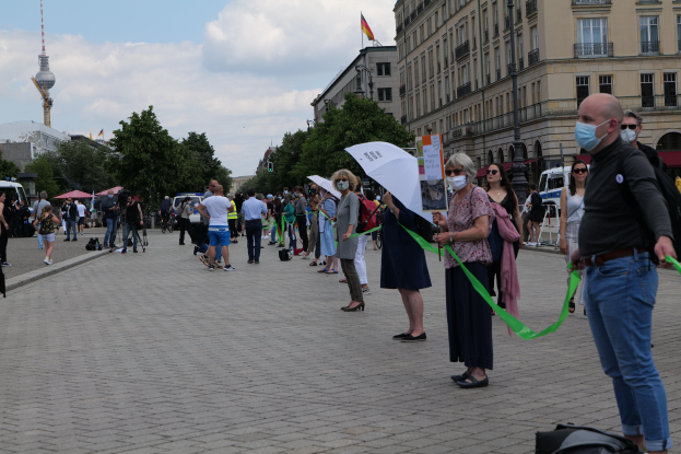 Eine Gruppe von Menschen marschiert auf einer Straße in Berlin, Deutschland, mit Schildern und Fahnen, einige tragen Masken und halten Schirme, mit Bäumen, Gebäuden, geparkten Fahrzeugen und einem Flaggenturm im Hintergrund bei bewölktem Himmel.