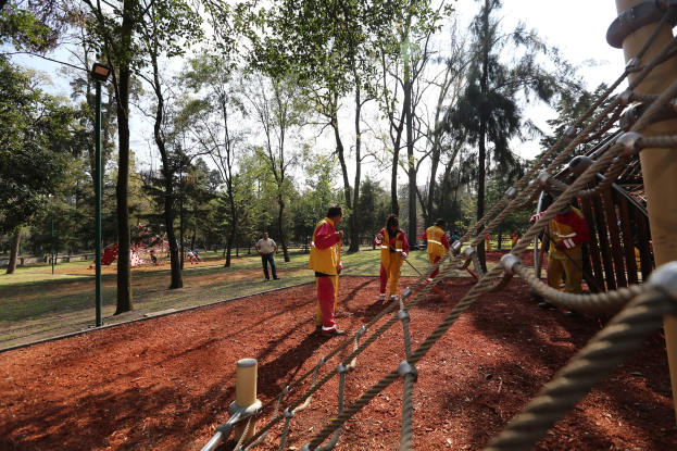 Gruppe von Menschen, die einen Seilparcours in einem Park mit Bäumen und erdbedecktem Boden durchläuft, mit einem Pfahl mit Seilen auf der rechten Seite.