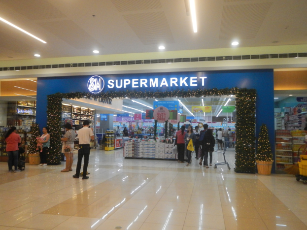 Menschen vor einem Supermarkt mit Einkaufstaschen, einem Einkaufswagen, Topfpflanzen, Regalen, Schildern und Deckenleuchten.