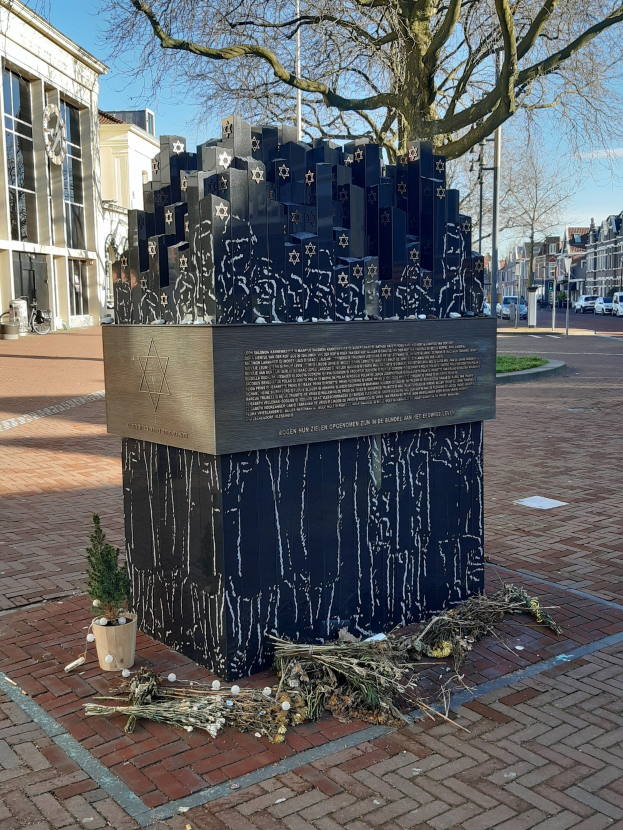 Denkmal in einem Stadtplatz für Holocaust-Opfer, mit einem Baum, Topfpflanze, verstreuten trockenen Blättern und umliegenden Gebäuden, Fahrzeugen, Polen und Bäumen.