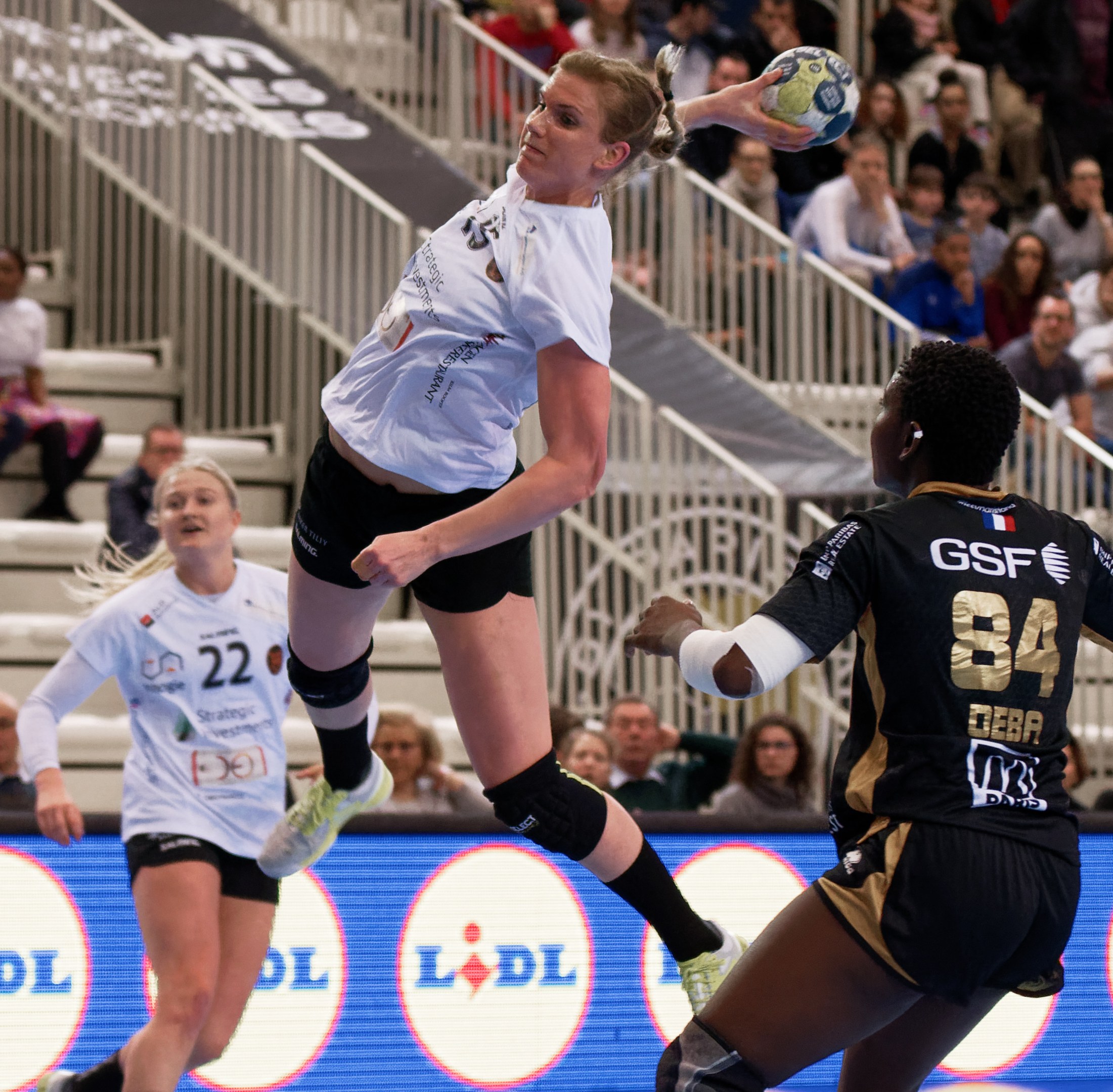 Eine Gruppe von Frauen, die auf einem Platz Handball spielen, mit einem Ball in der Luft, Zuschauern im Hintergrund und einer "Handball-Weltmeisterschaft 2019 - Tag 2"-Tafel.