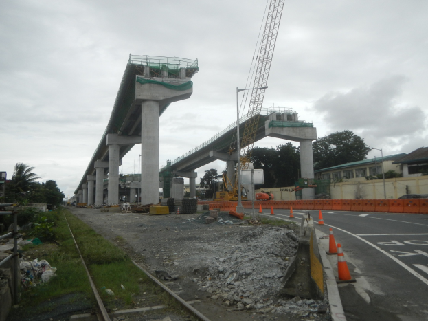 Baustelle mit einer Brücke im Hintergrund, Straße mit Absperrgittern markiert, verstreute Steine und Gras, Bahnschiene links, Bäume und Gebäude säumen die Straße und ein bewölkter Himmel.