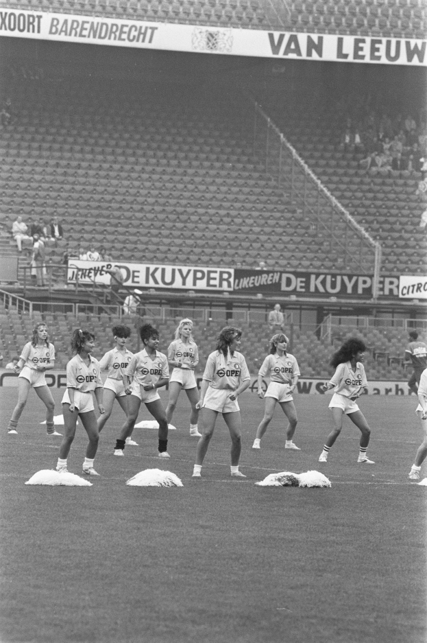 Schwarzes Bild einer Gruppe von Frauen auf einem Fußballfeld mit Zuschauern auf den Rängen und Bannern im Hintergrund.
