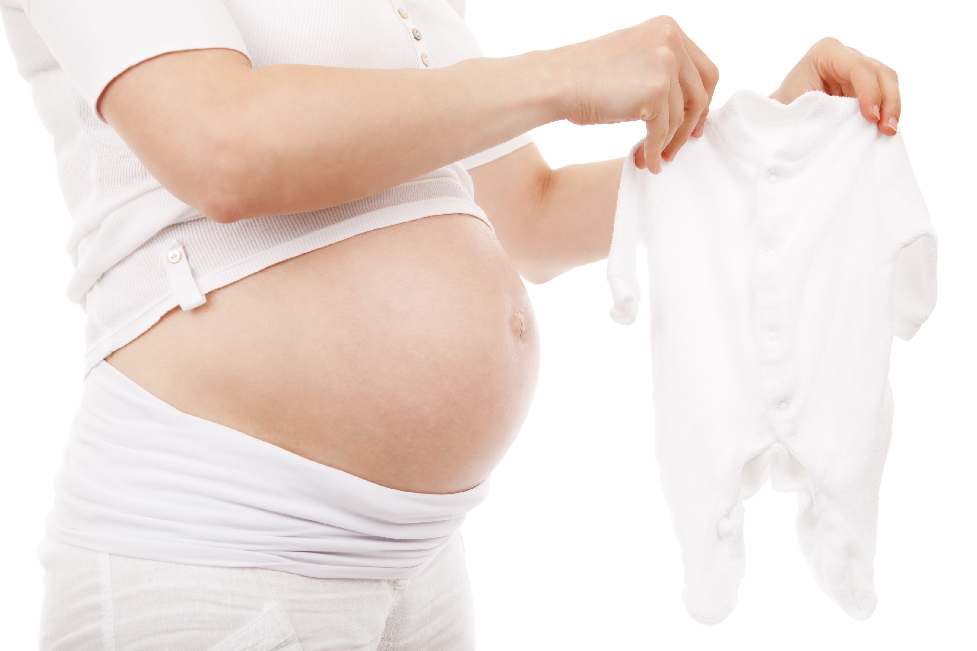 Schwangere Frau in einem weißen Kleid, die ein weißes Baby-Bodysuit hält und zufrieden lächelt.