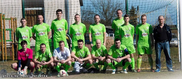 A group of men in green t-shirts, some squatting, stand on a soccer field with a ball and goal post, with trees, buildings, and vehicles in the background.