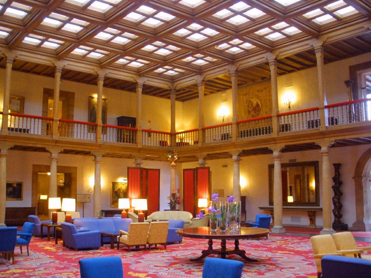 Ein geräumiger Hotellobby mit blauen Stühlen, Sofas, Tischen, Blumenvasen, Lampen, Säulen, Geländern, Lichtern, gerahmten Wandbildern, Fenstern mit Vorhängen und einem Teppichboden.