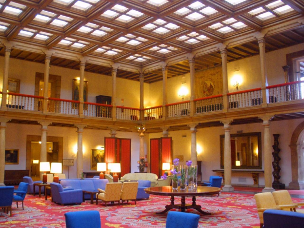 Ein geräumiger Hotellobby mit blauen Stühlen, Sofas, Tischen, Blumenvasen, Lampen, Säulen, Geländern, Lichtern, gerahmten Wandbildern, Fenstern mit Vorhängen und einem Teppichboden.