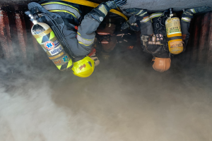 Eine Gruppe von Feuerwehrmännern in Helmen, Handschuhen und Sauerstoffflaschen arbeitet daran, ein Feuer in einem Tunnel zu löschen, wobei Rauch aus der Unterseite aufsteigt.