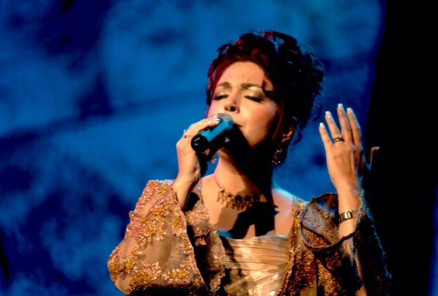 A woman in a gold dress singing into a microphone on stage at the Eurovision Song Contest with a blue and black background.
