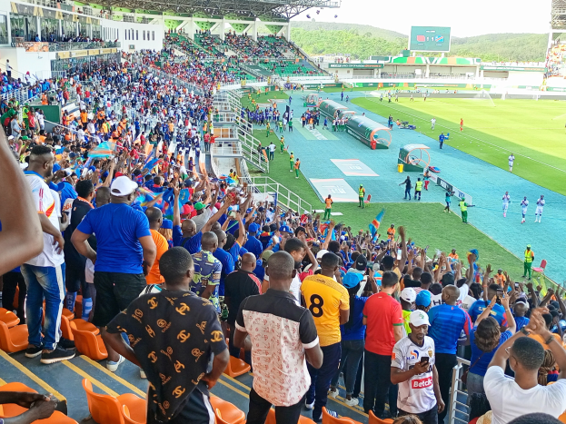Eine große Menschenmenge versammelt sich vor einem eingezäunten Fußballfeld, wobei einige auf Stühlen sitzen und andere stehen, während Hügel und ein klarer blauer Himmel die Kulisse bilden während der African Cup of Nations 2019.