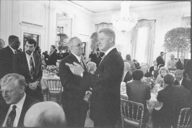 Schwarzes und weißes Foto von Präsident John F. Kennedy und Vizepräsident Lyndon B. Johnson, die sich die Hand schütteln, im Oval Office des Weißen Hauses umgeben von sitzenden und stehenden Personen, mit Blumenvasen, Gläsern und Tellern auf den Tischen.
