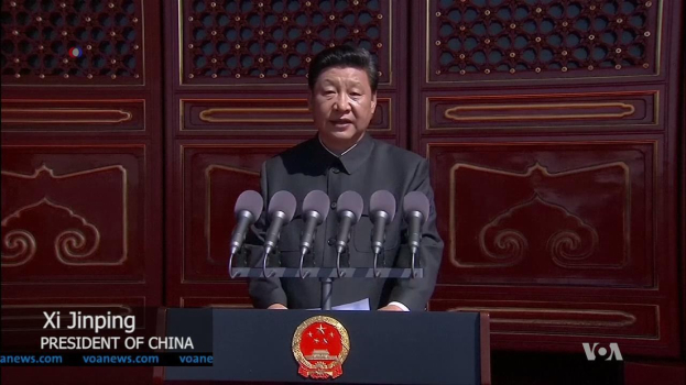 Xi Jinping steht an einem Pult mit Mikrofonen und Logo, hält eine Rede in der Großen Halle des Volkes in Peking, mit einer hölzernen Wand im Hintergrund.