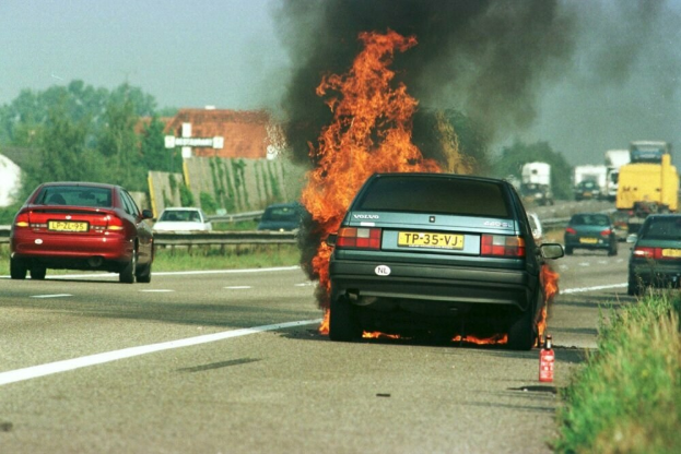 Ein Auto in Flammen auf der Stra├če, umgeben von anderen Fahrzeugen, mit B├Ąumen, Geb├Ąuden und einem klaren blauen Himmel im Hintergrund und einem Feuerl├Âscher auf der rechten Seite.