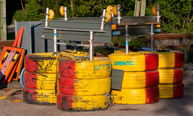 Ein Haufen gelber und roter Reifen auf einem Schotterfeld, umgeben von verschiedenen Gegenständen, mit Bäumen, Pflanzen und einem Schuppen im Hintergrund.