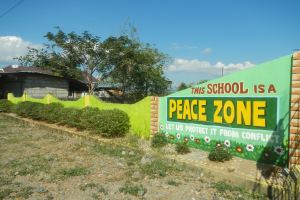 Eine Wand mit einem Schild, auf dem "Diese Schule ist eine Friedenszone" steht, umgeben von Pflanzen, Gras, Steinen, Bäumen, Häusern mit Dächern und Fenstern, einem Mast mit Drähten und einem bewölkten Himmel.