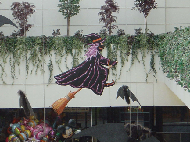 Halloween-Umzug mit einer Hexe auf einem Besen vor einem Gebäude, mit Spielzeug und Luftballons im Vordergrund, Efeu an einer Wand im Hintergrund und einem Geländer auf der rechten Seite.