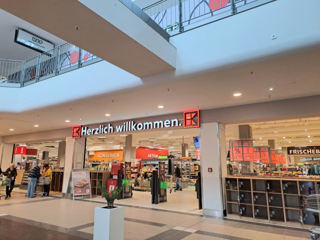 Das Innere eines Einkaufszentrums mit Menschen, die herumlaufen, Geschäften, Schildern mit Text, Lampen, Geländern und anderen Gegenständen, mit einer Topfpflanze unten und dem Text "Herzlich willkommen" auf den Schildern.