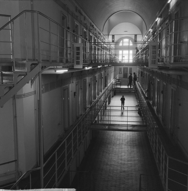 Schwarzes und weißes Foto eines Mannes, der einen langen Gefängnishofgang mit Gittern, Türen und Lampen sowie einem Fenster im Hintergrund entlanggeht.