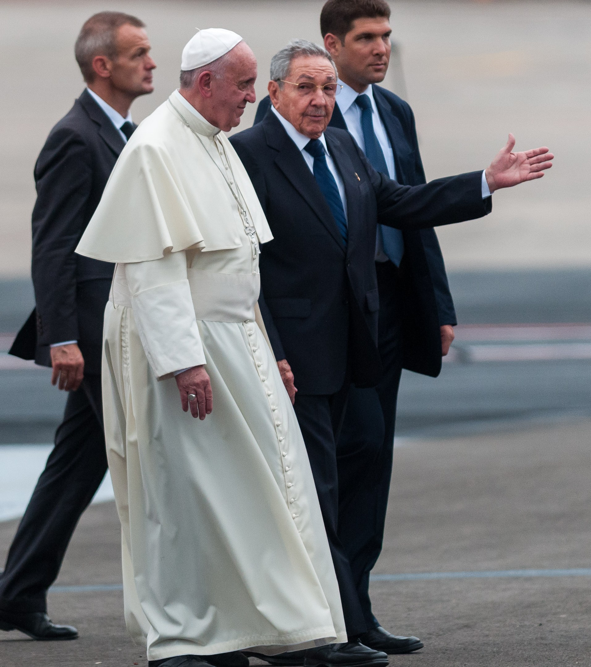 Pope Francis in a white robe and cap, accompanied by four men in black suits and ties, arriving at an airport in St. Peter's Square, Vatican City.