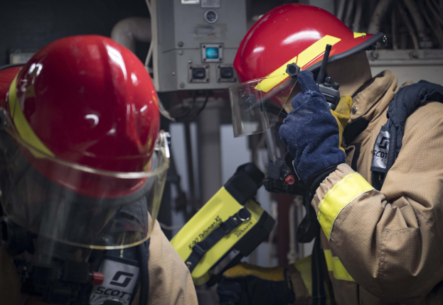 Zwei Feuerwehrleute in Schutzausrüstung arbeiten an einem Feuerhydranten während einer übungsstunde mit Maschinen und Kabeln im Hintergrund.