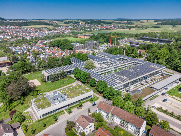 Luftaufnahme eines großen Gebäudes mit Solarmodulen auf dem Dach, umgeben von Häusern, Bäumen und Fahrzeugen, mit Hügeln und einem klaren blauen Himmel im Hintergrund.