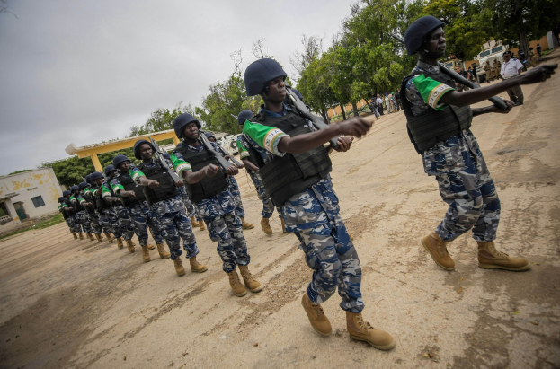 Nigerian soldiers march in formation down a dirt road, carrying rifles, with trees, buildings, and a vehicle visible in the background under a clear sky.