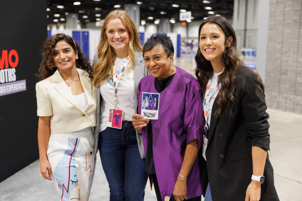 Vier Frauen mit ID-Karten stehen zusammen lachend vor einem Stand, mit einer Texttafel links daneben und Lichtern im Hintergrund.