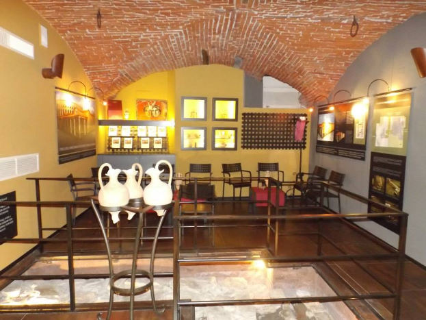 Ein Museumsraum voller Tische, Stühle, einem Geländer, Vasen, Texttafeln, Wandrahmen, Lampen und verschiedenen Ausstellungsstücken.