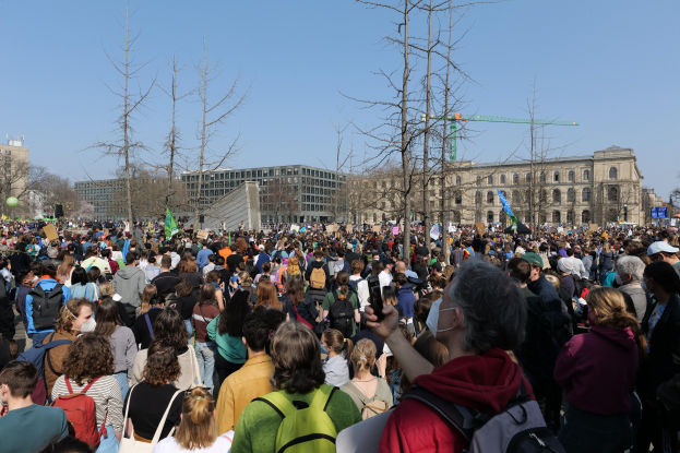 Eine große Menge Menschen mit Schildern und Taschen vor einem Gebäude mit Fenstern, Bäumen und einem klaren Himmel im Hintergrund, was auf eine Klimaprotest hingedeutet.