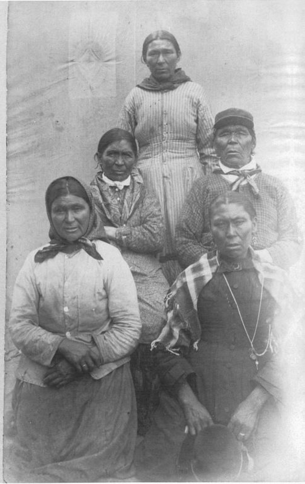 Schwarüß-weißes Foto einer Gruppe von indigenen Frauen, die in einer Reihe vor einer schlichten Wand stehen, einige tragen traditionelle Kleidung und Kopfschmuck.