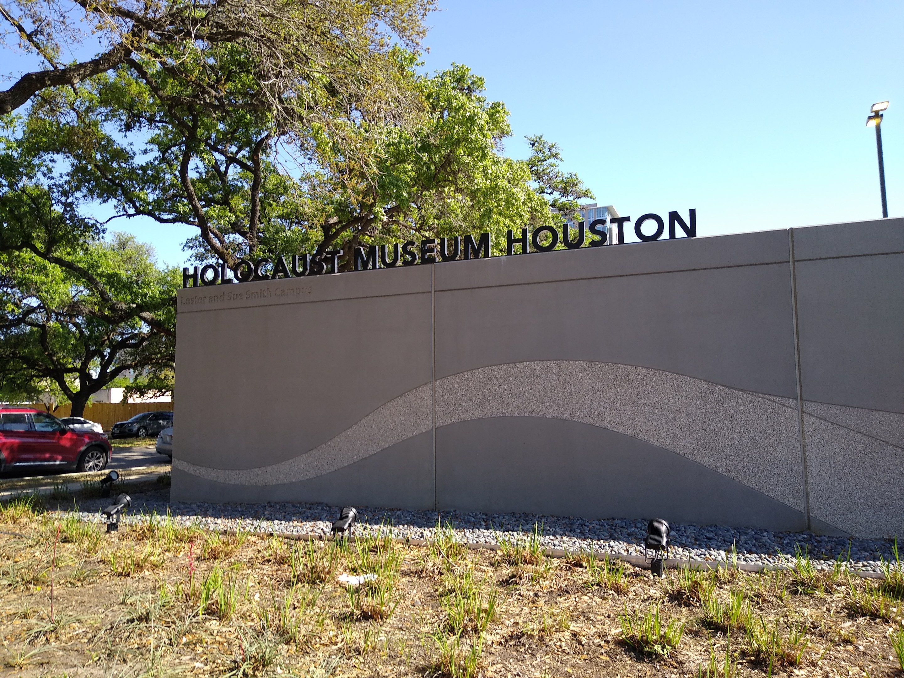 Exterior view of the Houston Holocaust Museum with engraved text wall, scattered stones, greenery, trees, passing vehicles, a street pole, and a cloudy sky.