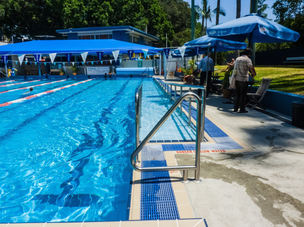 Großer Freibadpool mit schwimmenden Menschen, umgeben von Geländern, Sonnenschirmen, Stühlen, Tischen, Grünpflanzen und einem Gebäude unter einem klaren blauen Himmel.