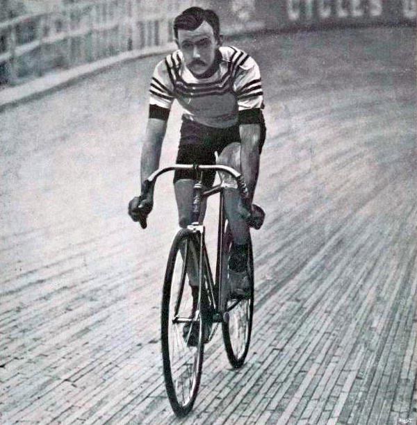 Schwarz-weiß-Foto eines Mannes, der auf einer Bahn mit einem Zaun und einer Tafel im Hintergrund Fahrrad fährt.