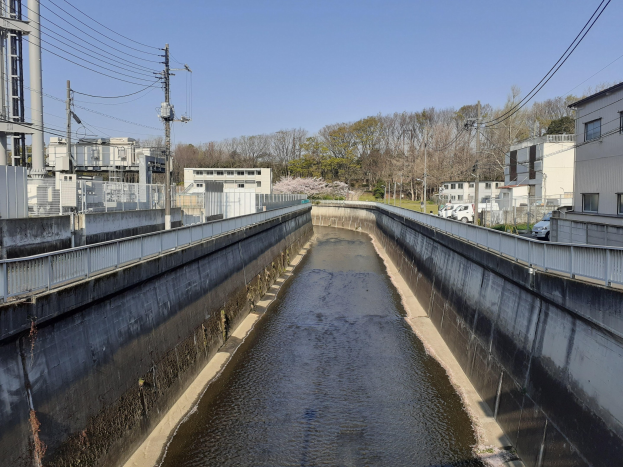 Small canal running through a city with buildings on either side, electric poles with wires, vehicles on the road, trees in the background, and a clear blue sky.