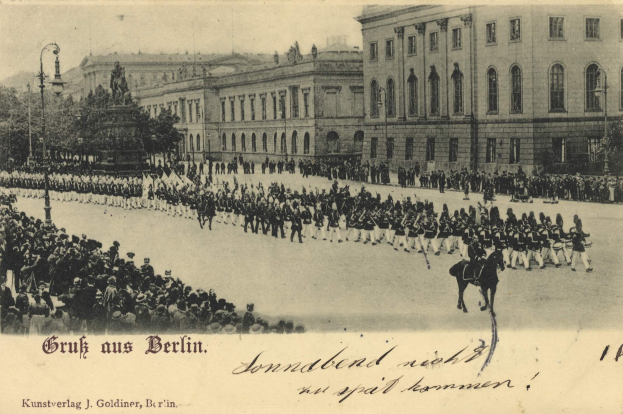 Ein altes Schwarz-Weiß-Foto eines Umzugs in Berlin, Deutschland, mit Menschen auf dem Boden und auf Pferden, einer Statue auf einem Sockel, Straßenlaternen, Bäumen, Gebäuden mit Fenstern und einem bewölkten Himmel, mit Text am unteren Rand des Bildes.