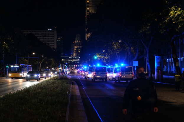 Eine Stadtstraße bei Nacht mit Polizeiwagen, Bussen, Fußgängern auf dem Gehweg, Gras, Bäumen, Gebäuden und beleuchteten Laternen.