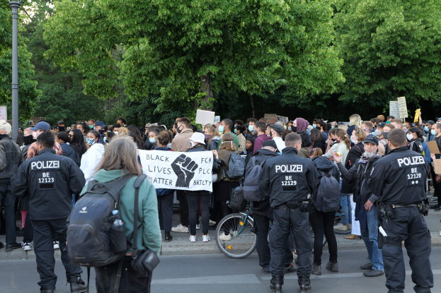 Eine große Gruppe von Menschen nimmt an einer Black Lives Matter Demonstration in Berlin teil, einige halten Schilder und tragen Kappen und Taschen, während im Vordergrund ein Fahrrad und im Hintergrund Bäume und ein Mast zu sehen sind.