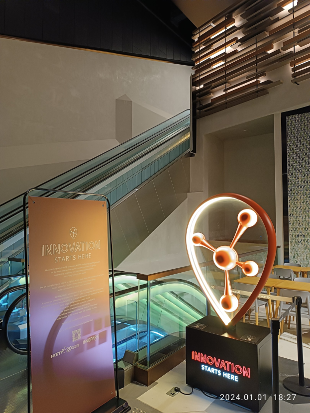 Großes Schild in einem Gebäude mit einer Rolltreppe im Hintergrund, Tische und Stühle auf der rechten Seite und eine Tafel mit "Innovation Starts Here" auf der linken Seite, die auf die HSTP20 Expo hinweist.