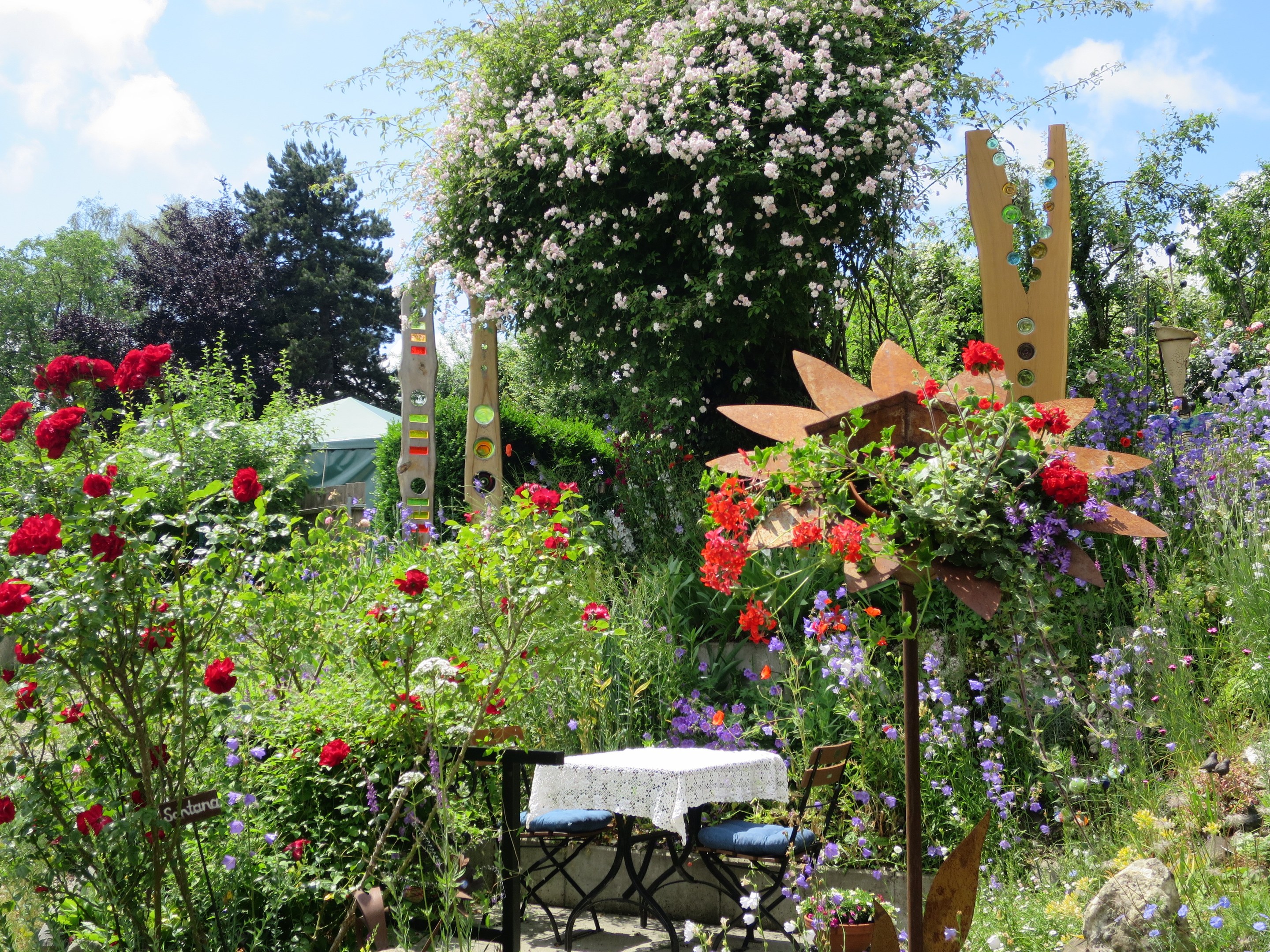 Ein Garten auf der Chelsea Flower Show mit bunten Blumen, Pflanzen, einem Tisch und Stühlen in der Mitte, einem Tuch auf dem Tisch und Bäumen im Hintergrund unter einem bewölkten Himmel.