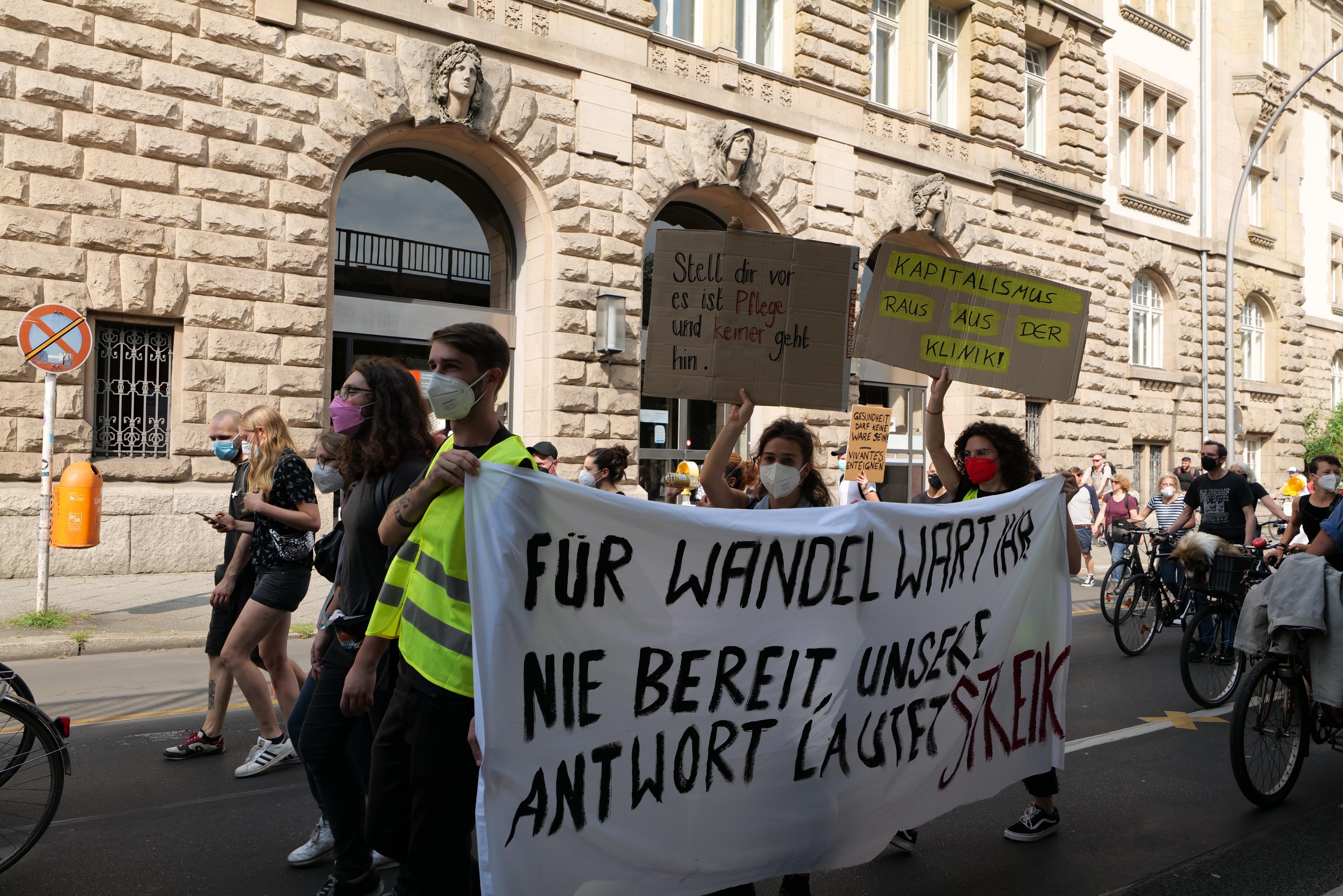 A protest march in Berlin with people holding signs and riding bicycles, passing a building with arched windows, pillars, and sculptures, and trees in the background.