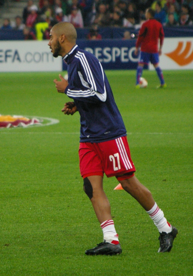 Ein professioneller Fußballspieler in einem blauen T-Shirt und roten Shorts läuft auf einem Feld, während im Hintergrund eine Gruppe von Menschen sitzt und Werbetafeln zu sehen sind.