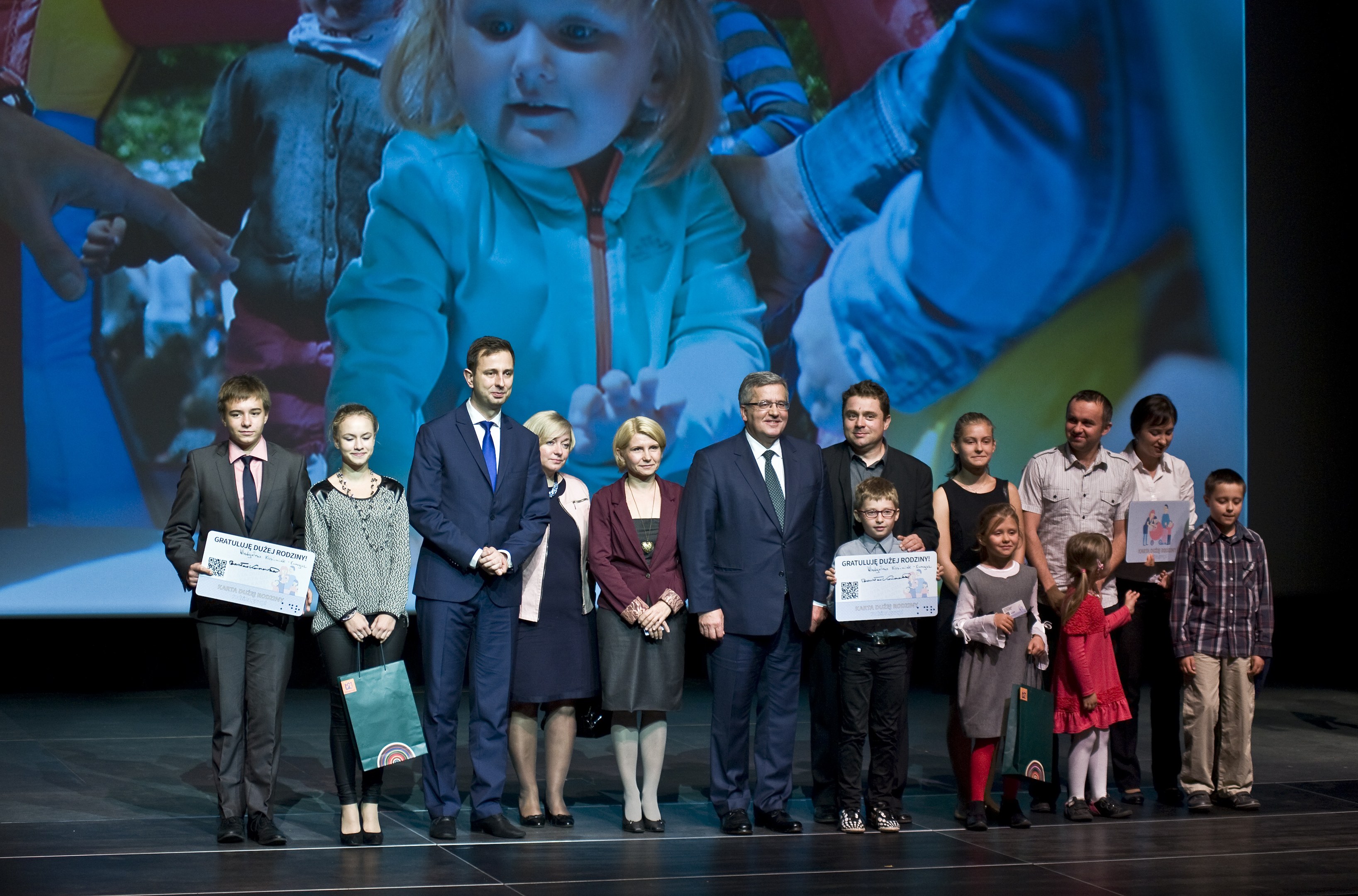 Gruppe von Menschen auf einer Bühne mit Taschen und Urkunden, im Hintergrund ein Bildschirm mit einem Kind und anderen Personen