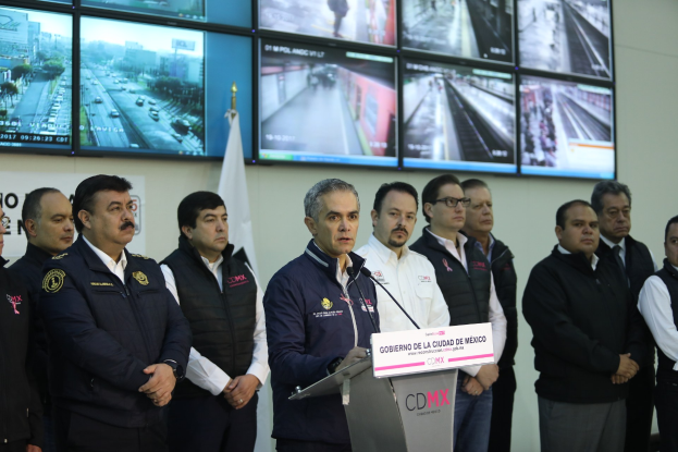 Eine Gruppe von Männern vor einem Rednerpult mit einem Mikrofon und einer Tafel mit der Aufschrift "Mexikanische Zoll- und Grenzschutzbehörde", mit Fernsehgeräten und einer Flagge an der Wand dahinter.