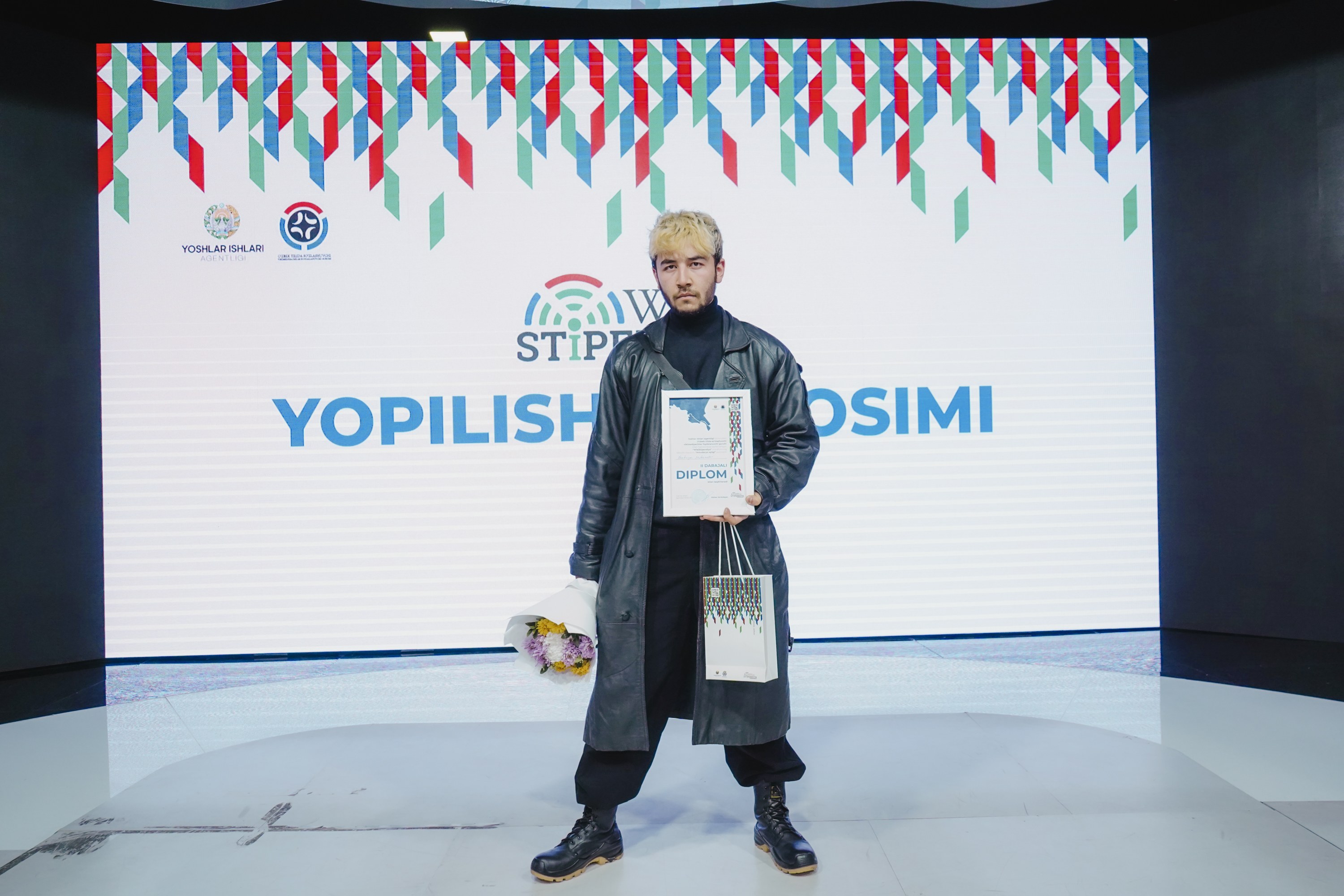 A man on stage holds a bouquet and a certificate while a banner reading "Yopilish Cosimi 2019" is visible behind him.