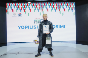 A man on stage holds a bouquet and a certificate while a banner reading "Yopilish Cosimi 2019" is visible behind him.