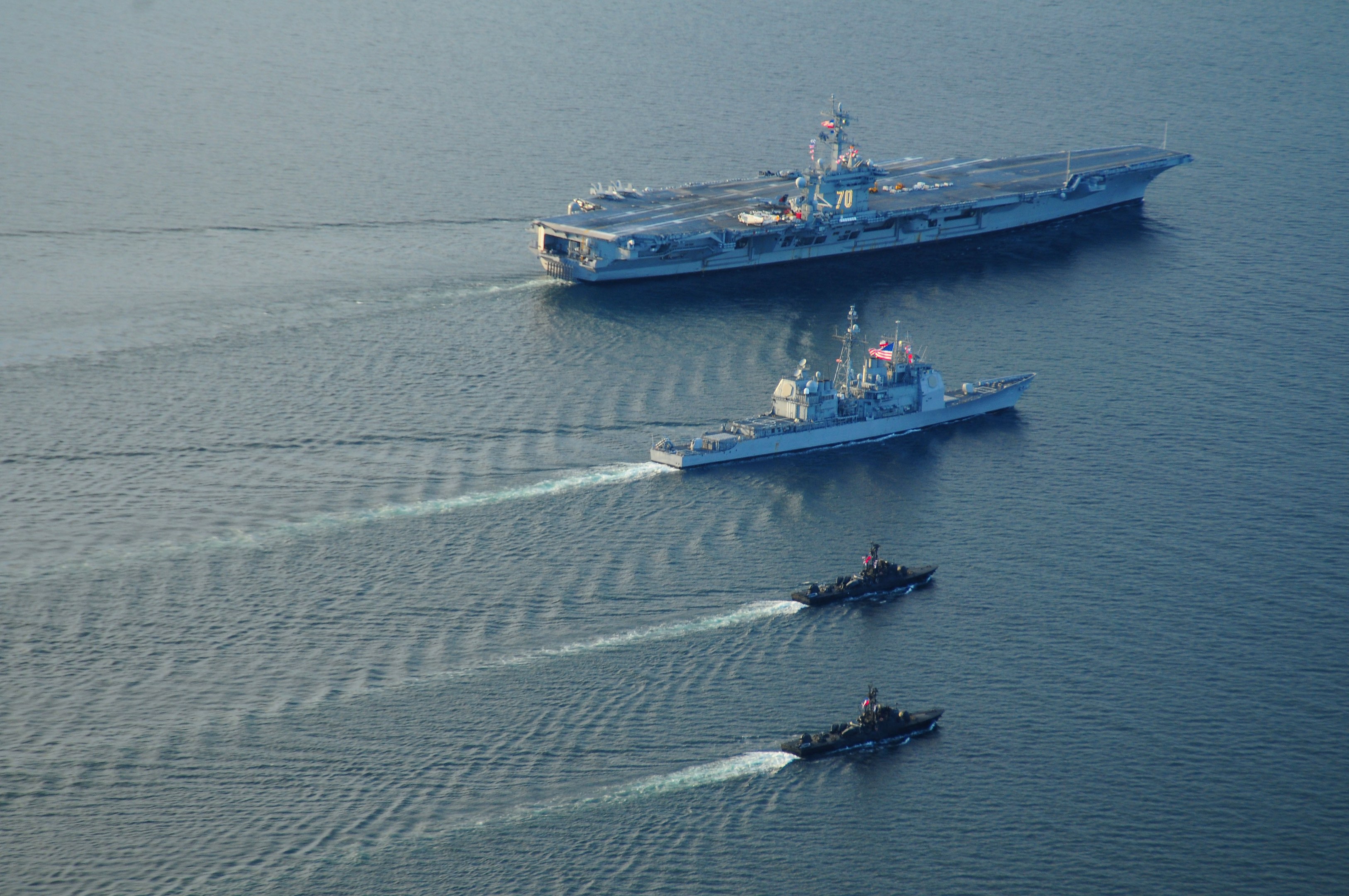 A large aircraft carrier, the USS George H W Bush CVN-68, sails through the Strait of Hormuz surrounded by two small boats, with flags and text visible on the vessel.