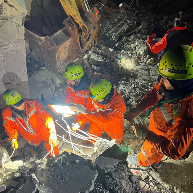 Gruppe von Menschen in orangefarbenen Uniformen und grünen Helmen bei der Arbeit an Schutt mit einem Kran im Hintergrund bei einem Einsatz nach einem Einsturz.