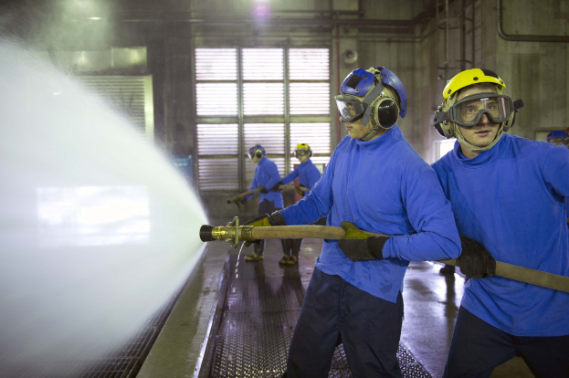 Gruppe von Männern in blauen Hemden und gelben Helmen, die an einer Maschine arbeiten, während einer das Wasser auf den Boden sprüht, in einer Fabrikumgebung mit sichtbaren Wänden, Fenstern, Rohren und Lampen.