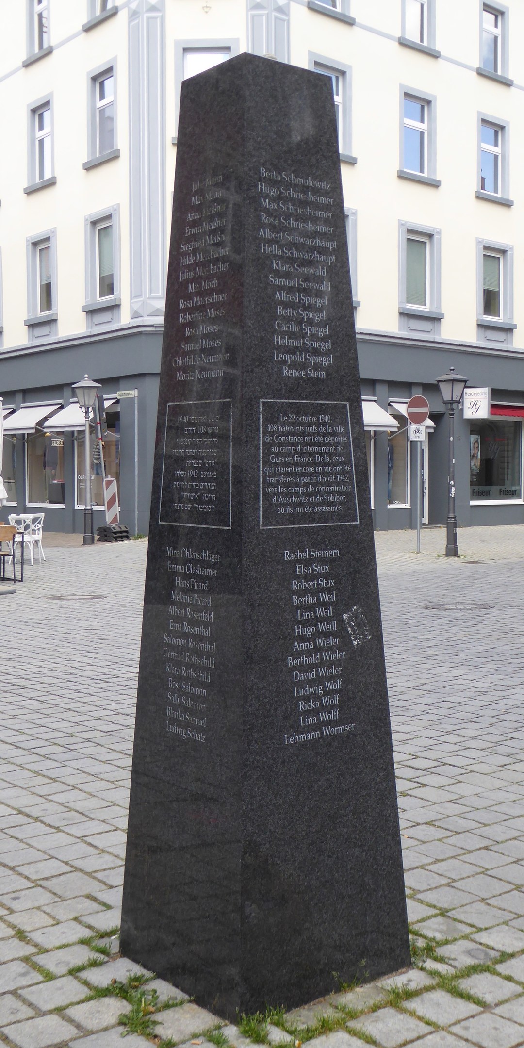 Ein hohes Denkmal mit eingravierter Schrift steht auf einem Platz, umgeben von Gebäuden, Straßenmöbeln und Menschen, mit dem Himmel im Hintergrund.